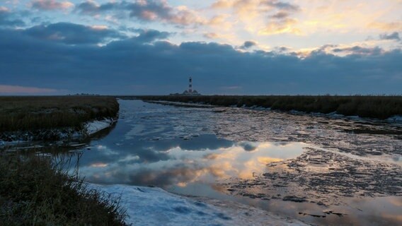 Im Vordergrund ist eine leichte Eisschicht auf dem Wasser zu sehen, das zwischen Dühnengras zum Westerhever Leuchtturm führt. © Wenke Stahlbock Foto: Wenke Stahlbock