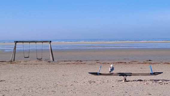 Die Nordsee am Horizont, im Vordergrund Strand mit einer Schaukel zur Linken und einer Wippe zur Rechten. Auf der Wippe sitzt eine Möwe. © Michael Fugmann Foto: Michael Fugmann