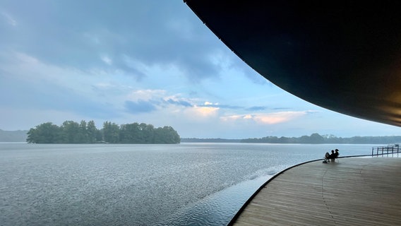 Zwei Personen sitzen unter der Eutiner Seebühne und schauen auf das Wasser. Währenddessen fällt leichter Regen. © Roland Schulenberg Foto: Roland Schulenberg