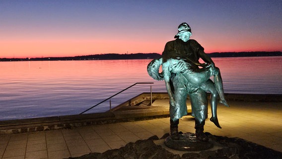 Die Statue "Gerettet", die einen Fischer zeigt, der eine Frau vor dem Ertrinken rettet, vor dem Kieler Hafen bei Abendhimmel in Heikendorf. © Ottmar Schulz Foto: Ottmar Schulz