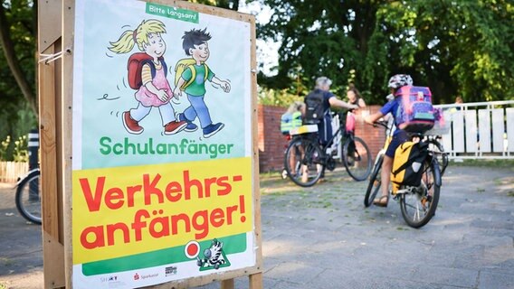 Im Vordergrund ist ein Hinweisschild mit der Aufschrift "Schulanfänger, Verkehrsanfänger zu sehen", welches die Autofahrer auf die neuen Schulkinder aufmerksam machen soll. Im Hintergrund befinden sich Kinder auf Fahrrädern, die ihren Ranzen auf dem Rücken tragen. © dpa Foto: Christian Charisius/dpa