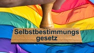 Ein symbolischer Holzstempel mit der Aufschrift "Selbstbestimmungsgesetz", gehalten von einer Hand im Anschnitt vor einer, im Wind wehenden Regenbogenfahne im Hintergrund. © picture alliance / SULUPRESS.DE Foto: Torsten Sukrow / SULUPRESS.DE