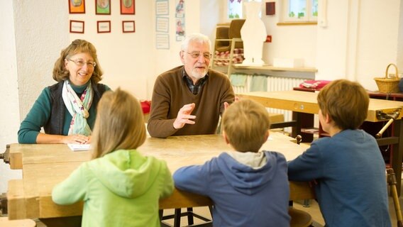 Zwei Senioren sitzen drei Kindern in einem Klassenzimmer gegenüber und unterstützen sie bei ihren Schulaufgaben, indem sie ihnen etwas erklären. © picture alliance / dpa Themendienst Foto: Tobias Hase