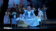 Auf einer Bühne steht ein Bett, auf dem Bett sitzt eine Person, wetere flankieren die Szene aus einer Oper © NDR Foto: NDR Screenshot