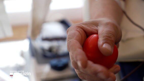 Eine Hand drückt auf einem Anti-Stress-Ball, im Hintergrund ist ein sicher füllender Beutel einer Blutspende auf einem Gerät. © NDR Foto: NDR Screenshot
