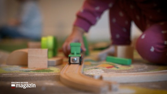 Ein Kind spielt mit einer Holzeisenbahn auf einem Spieleteppich © NDR Foto: NDR Screenshot