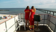 Zwei Rettungsschwimmerinnen der DLRG blicken von einem Turm aus auf den Strand hinab © NDR Foto: NDR Screenshot