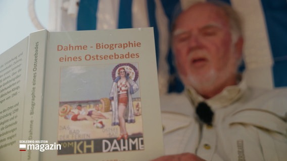 Ein Mann in einem Strandkorb hält ein Buch in die Kamera, der Titel des Buches ist: "Dahme - Biographie eines Ostseebades" © NDR Foto: NDR Screenshot