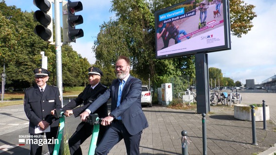 Schleswig-Holstein Verkehrsminister Claus Ruhe Madsen steht mit zwei Polizisten vor einer Werbetafel die für sicheren Umgang mit E-Rollern wirbt © NDR Foto: NDR Screenshot