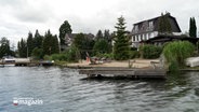 Eine Blick vom Wasser auf ein Holz mit einem Spielplatz und einem Steg davor © NDR Foto: NDR Screenshot