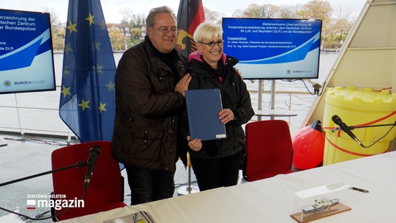 Zwei Personen posieren mit einer Mappe vor einigen Fahnen © NDR Foto: NDR Screenshot