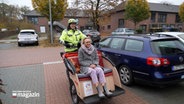 Eine Frau wird von einem Mann in einer Rikscha über einen Parkplatz gefahren © NDR Foto: NDR Screenshot