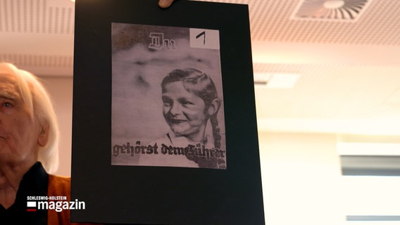 Ein mann hält bei einem Vortrag ein Plakat aus der Hitler-Zeit empor. Darauf ein Mädchen udn der Schriftzug "Du gehörst dem Führer" © NDR Foto: NDR Screenshot