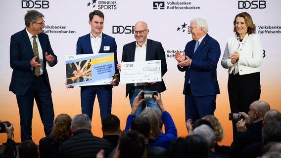 Bundespräsident Frank-Walter Steinmeier (2.v.r.) zeichnet zusammen mit Thomas Weikert (l), Präsident des Deutschen Olympischen Sportbundes (DOSB), und Marija Kolak (r), Präsidentin des Bundesverbandes der Deutschen Volksbanken und Raiffeisenbanken (BVR), bei der Verleihung der "Sterne des Sports" in Gold den Verein TSB Flensburg v. 1865 e.V. (Schleswig-Holstein) mit dem ersten Platz beim "Stern des Sports in Gold 2025" aus. © picture alliance/dpa Foto: Bernd von Jutrczenka