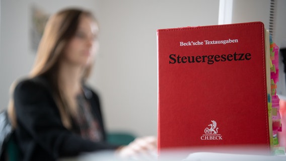 Eine Frau arbeitet an einem Comupterarbeitsplatz, im Vordergrund ist ein Steuergesetzbuch zu sehen. © Picture Alliance Foto: Marijan Murat