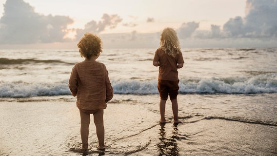 Zwei Kinder stehen knöchelhoch im Wasser an einem Strand bei Sonnenuntergang. © IMAGO / Cavan Images 