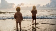 Zwei Kinder stehen knöchelhoch im Wasser an einem Strand bei Sonnenuntergang. © IMAGO / Cavan Images 