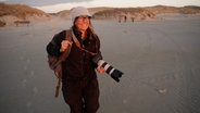 Eine Frau geht mit einer Kamera in der Hand am Strand entlang. © NDR 