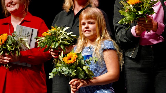 Ein Mädchen steht auf einer Bühne, es hält einen Blumenstrauß in der Hand © NDR Foto: NDR Screenshot