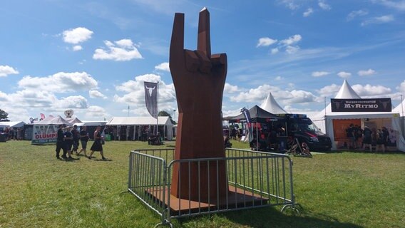 Ein riesiges Monument aus rotem Stahl steht auf dem Rasen, es zeigt eine Hand, die den Zeigefinger und kleinen Finger nach oben streckt. © NDR Foto: Pauline Reinhardt