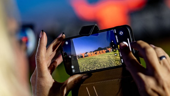 Wacken: Eine Besucherin des Wacken Open Air fotografiert mit ihrem Smartphone das Festival-Logo, welches in großen Metallbuchstaben auf einer Wiese steht. © dpa Foto: Axel Heimken