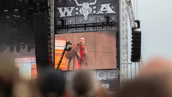 Ein Künstler in rotem Rollkragenpulli und Pelzmantel auf einem Bühnenmonitor. © NDR Foto: Dominik Dührsen
