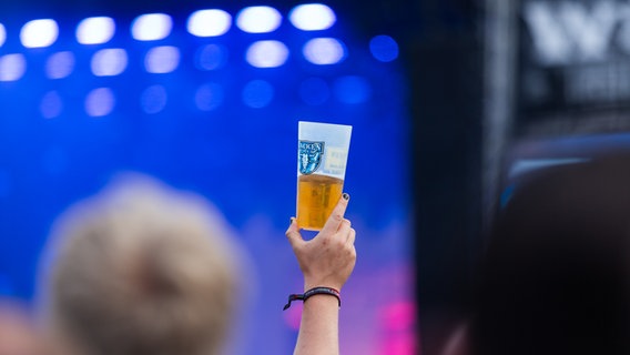 Eine Hand hält einen Plastikbecher mit Bier in einer Menschenmenge vor einer Konzertbühne. © NDR Foto: Dominik Dührsen