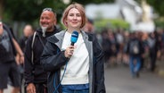 NDR Reporterin Vinietta Richter steht mit einem Mikrofon in der Hand auf einer belebten Straße in Wacken. © NDR Foto: Dominik Dührsen