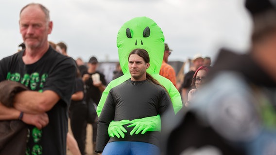 Ein langhaariger Mann trägt ein Alien-Kostüm und steht auf einem Festivalgelände. © NDR Foto: Dominik Dührsen