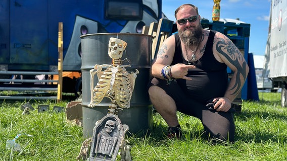 Ein Mann kniet auf dem Boden und macht den "Metalgruß" neben einem Model-Skelett © NDR Foto: Jörn Zahlmann