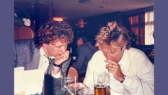 1987 in einer Kölner Kneipe: Uwe Bahn sitzt mit Rock-Legende Rod Stewart an einem Tisch, Stewart hält einen HSV-Wimpel in der Hand © Uwe Bahn 