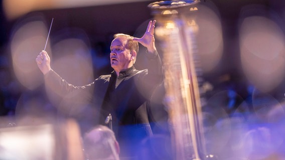 Dirigent Frank Strobel steht auf der Bühne mit beiden Armen erhoben, den Taktstock in der rechten Hand. Er schaut an der Kamera vorbei. © NDR Foto: Mischa Neugebauer