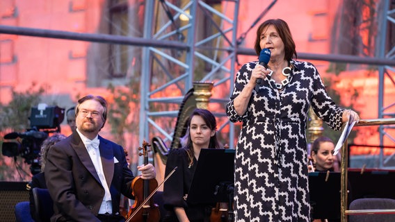 Eine Frau mit Mikrofon und Orchestermusiker:innen auf einer Bühne © NDR Foto: Helge Krückeberg