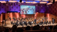 Konzert der Orchester-Detektive: Die NDR Radiophilharmonie, auf der Leinwand eine Schulklasse im Klassenzimmer © NDR Foto: Micha Neugebauer