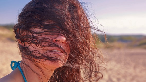 Eine Frau steht bei windigem Wetter an einem Strand. Ihre Haare bedecken ihr Gesicht. © Patrick Lohmüller / photocase.de Foto: Patrick Lohmüller