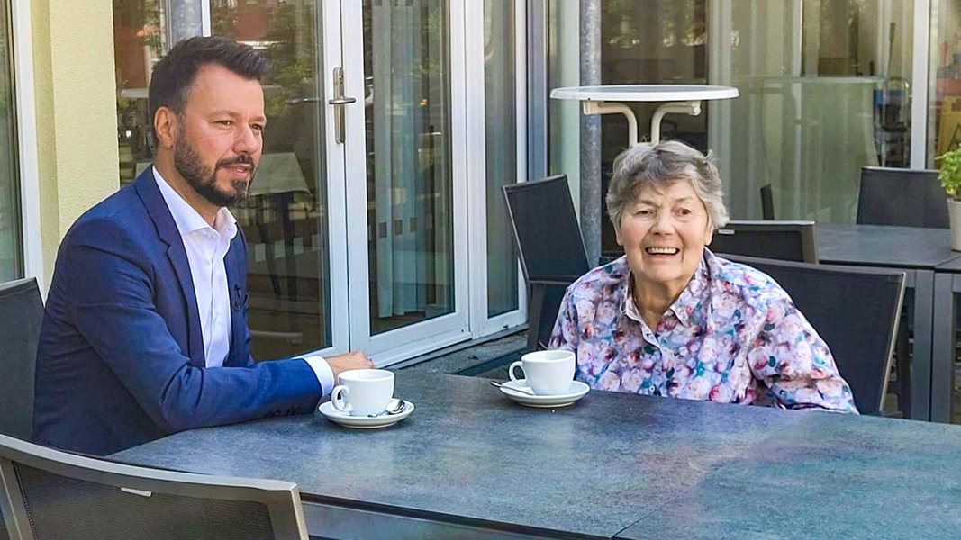 Heimleiter André Drummond und Bewohnerin Irmgard Eckert sitzen vor dem Gast- und Krankenhaus in Hamburg-Poppenbüttel an einem Tisch.