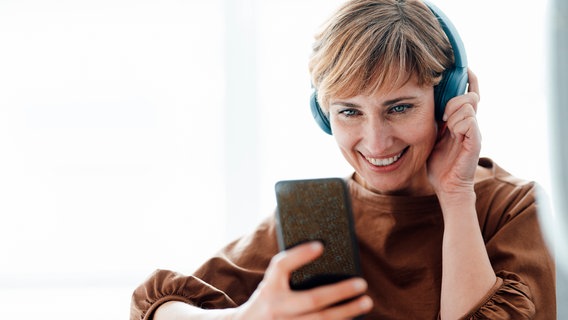 A woman wearing headphones smiles and looks at her smartphone.  © imago images Photo: Westend61