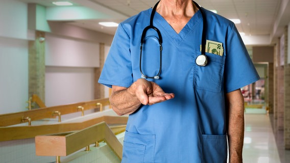 Arzt steht im Krankenhausflur und hält seine Hand ausgestreckt vor dem Körper, in der Brusttasche seines Arztkittels stecken Dollarscheine. © Colourbox 
