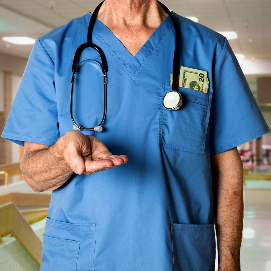 Arzt steht im Krankenhausflur und hält seine Hand ausgestreckt vor dem Körper, in der Brusttasche seines Arztkittels stecken Dollarscheine. © Colourbox 