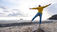 Ein Mann balanciert auf einem Felsbrocken am Strand. © Picture Alliance Foto: Uwe Umstätter