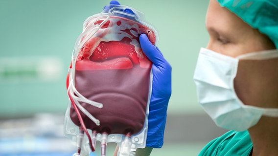 Eine Frau in OP-Kleidung hält eine Blutkonserve (durchsichtiger Beutel mit Blut) hoch © Friso Gentsch/dpa Foto: Friso Gentsch