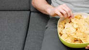 Ein Mann sitzt auf einem Sofa und greift in eine Schüssel voller Chips, die er auf dem Schoß hat © colourbox 