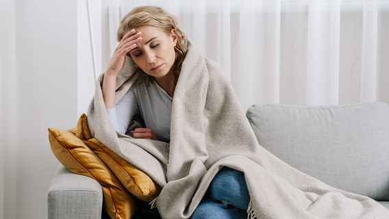 Eine Frau sitzt in eine Decke gewickelt erschöpft auf dem Sofa. © IMAGO / Dreamstime 