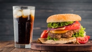 Ein Glas Cola mit Eiswürfeln steht auf einem Tisch. Daneben ein Tablett, auf dem ein Hamburger angerichtet ist. © IMAGO / Dreamstime Foto: IMAGO / Dreamstime