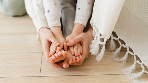 Hand-Mund-Fuß-Krankheit, Symbolbild: Hände und Füße von Mutter und Kind liegen übereinander. © IMAGO/ Westend61 Foto: Evgenia Sunegina