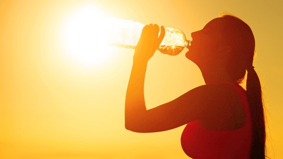 Aus einer großen Plastik-Wasserflasche trinkende, schlanke Frau als Silhouette im Gegenlicht vor einem sonnengelben Himmel. © Colourbox. 