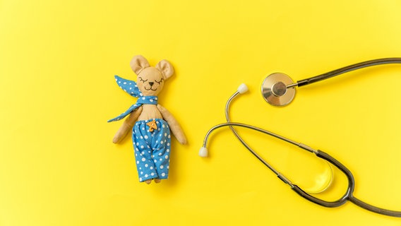 Draufsicht: Auf einer leuchtend gelben Flächen liegen ein schmaler Teddybär mit einer weiß gepunkteten blauen Hose und einem ebensolchen Halstuch und ein Stethoskop. © Colourbox.de 
