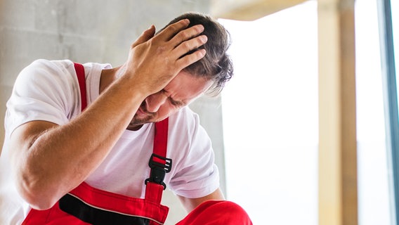 Handwerker sitzt nach Sturz auf den Kopf neben umgestürzter Leiter auf dem Boden und hält Hand an die Schläfe (Bild: colourbox.de) © colourbox.de 