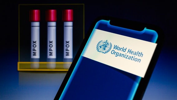 Das Logo der World Health Organization (WHO) ist auf einem Smartphone zu sehen, dahinter stehen Test-Röhrchen mit der Aufschrift "Mpox". © picture alliance / ZUMAPRESS.com | Andre M. Chang 