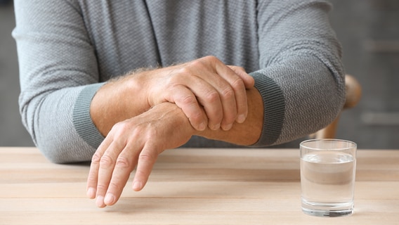 Ein Mann hält seine zitternde Hand, vor ihm ein Wasserglas. © IMAGO / Depositphotos 